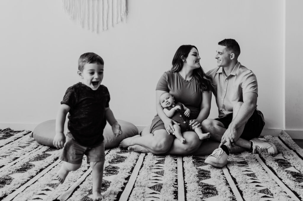Little boy running toward camera, which mom and dad gaze lovingly at each other with newborn baby. 